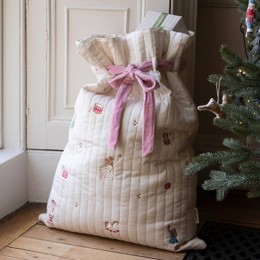 A quilted cream Christmas sack filled with gifts, tied with a pink ribbon, standing on a wooden floor next to a decorated Christmas tree.