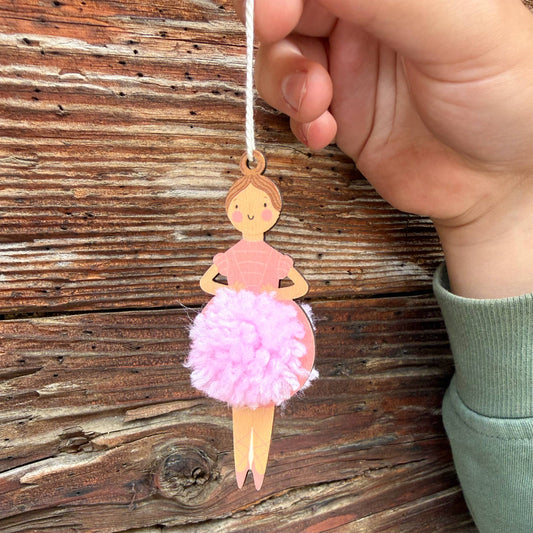 A hand holds a wooden ballerina ornament with a pink pom-pom skirt against a rustic wooden plank background.