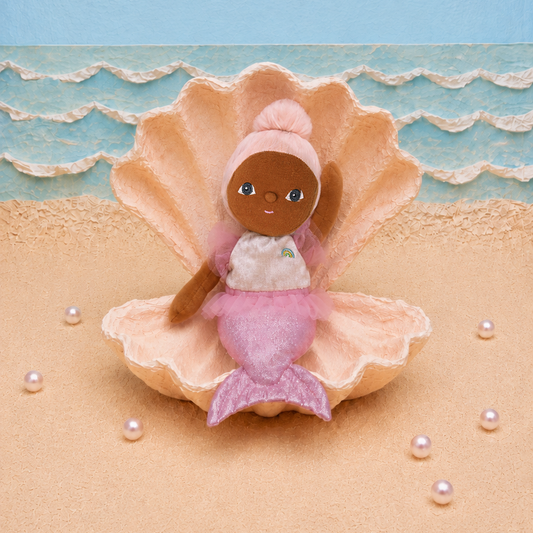 A small mermaid doll with brown skin, blue eyes, and pink hair sits inside a large peach seashell on a sandy beach set. The background features a light blue sky and ocean wave pattern, with scattered white faux pearls on the sand.