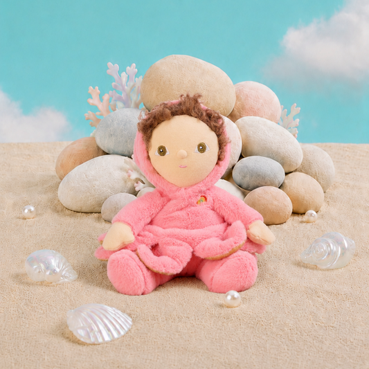 A plush Octopus doll with brown hair sits on a sandy surface in front of a pile of smooth stones, with small seashell props and a bright blue sky backdrop.