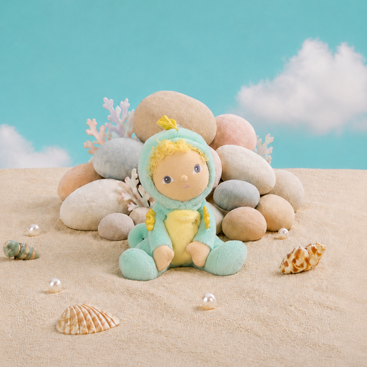 A plush Seahorse doll wearing a mint green and yellow hooded outfit sits on sand in front of a pile of smooth stones, with small seashells and coral-like decor against a turquoise sky backdrop with white clouds.