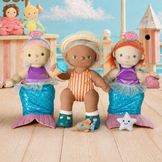 Three mermaid-themed dolls seated on a wooden deck with a beach and sky backdrop. The center doll wears an orange-and-white striped swimsuit and white hat, while the two side dolls wear purple tops and blue mermaid tails with pink fins.