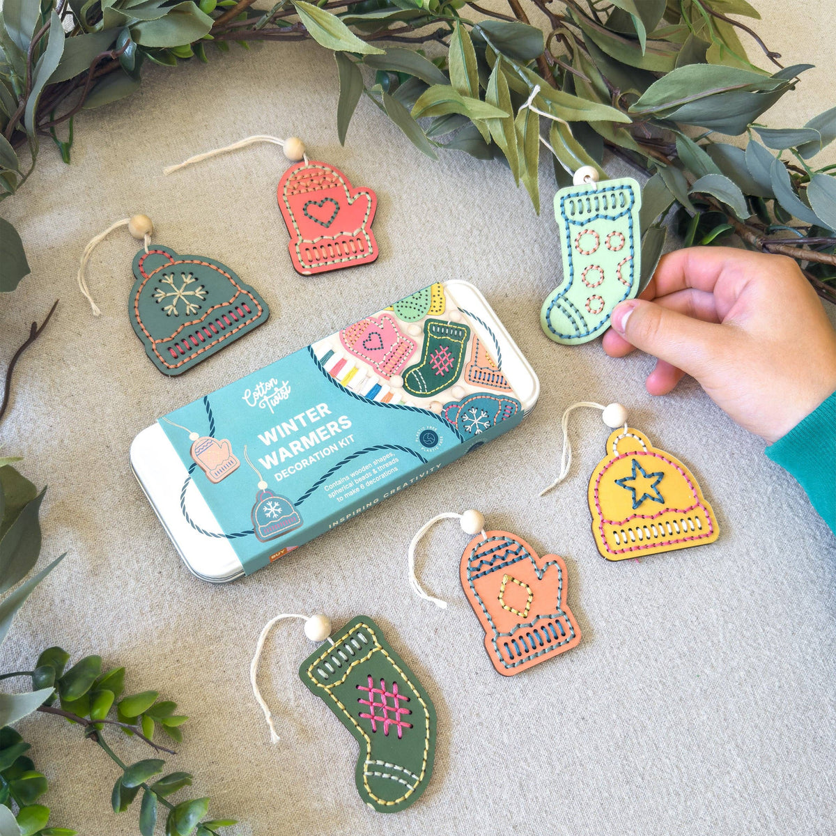 A tin of hand-embroidered wool felt Christmas ornaments is displayed on a neutral fabric surface with green foliage accents. A person's hand is holding one of the green stocking ornaments.