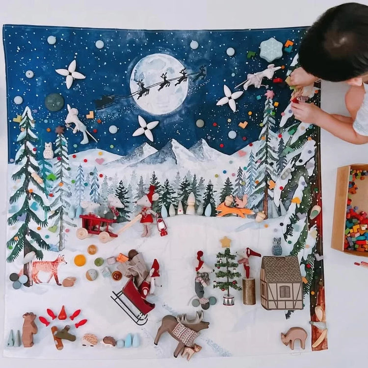 A child is actively building a large, colorful wooden Christmas scene with natural wood pieces on a Wonderie play cloth. Illustrating with Santa, reindeer, trees, mountains, and a moon, on a white background.