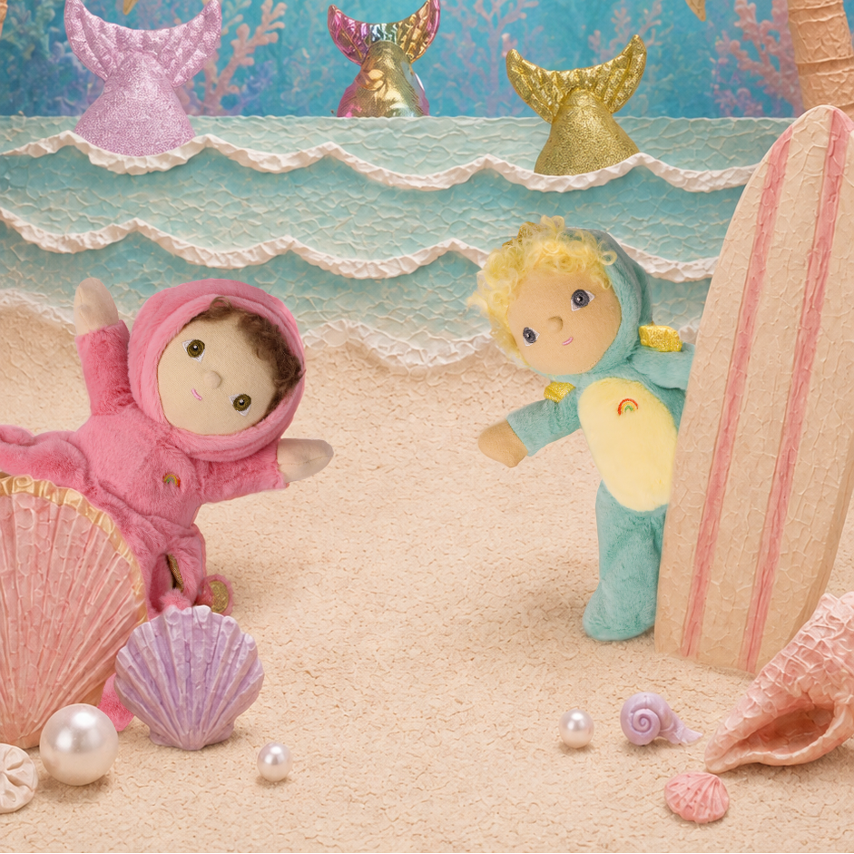 A whimsical underwater mermaid-themed scene with two small plush dolls dressed in pink and blue hooded outfits, surrounded by seashells, palm trees, and a striped surfboard against a blue watercolor backdrop.