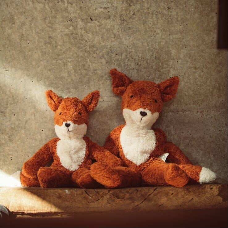Two fox plush toys sitting on a wooden surface with a textured beige wall background.
