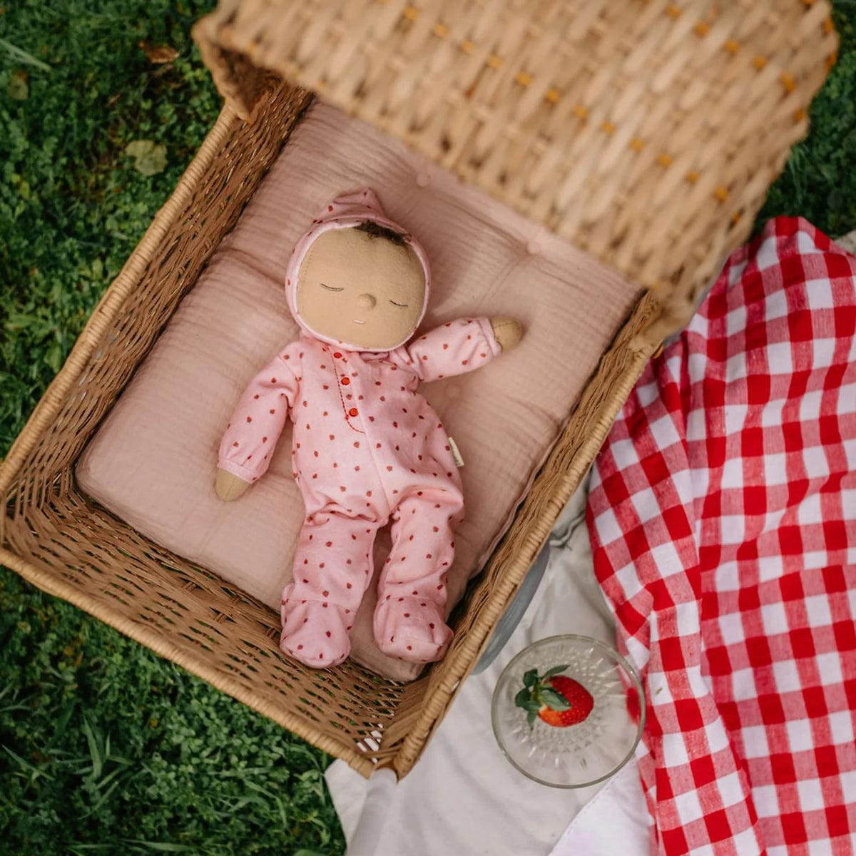 A dozy dinkum doll in a pink outfit lying in a wicker basket on grass with a red and white checkered blanket.
