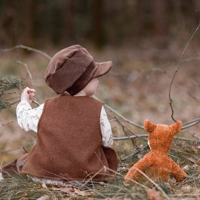 A child wearing a brown vest and matching hat sits outdoors in a wooded setting, facing away from the camera, with a small orange stuffed foxbeside them.