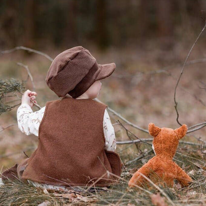 A child wearing a brown vest and matching hat sits outdoors in a wooded setting, facing away from the camera, with a small orange stuffed foxbeside them.