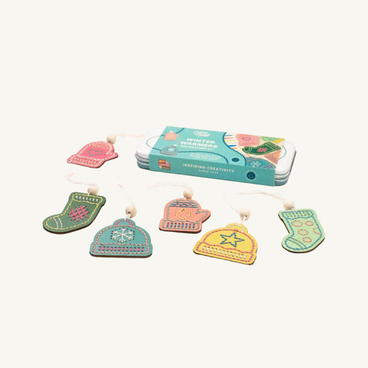 A set of colorful embroidered winter-themed ornaments shaped like a mitten, stocking, hat, and boot, displayed next to their teal packaging box on a light cream background.