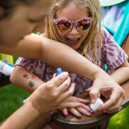 A young girl with pink patterned sunglasses smiles while her mother applies nail polish to her fingernails outdoors on a wooden bench.