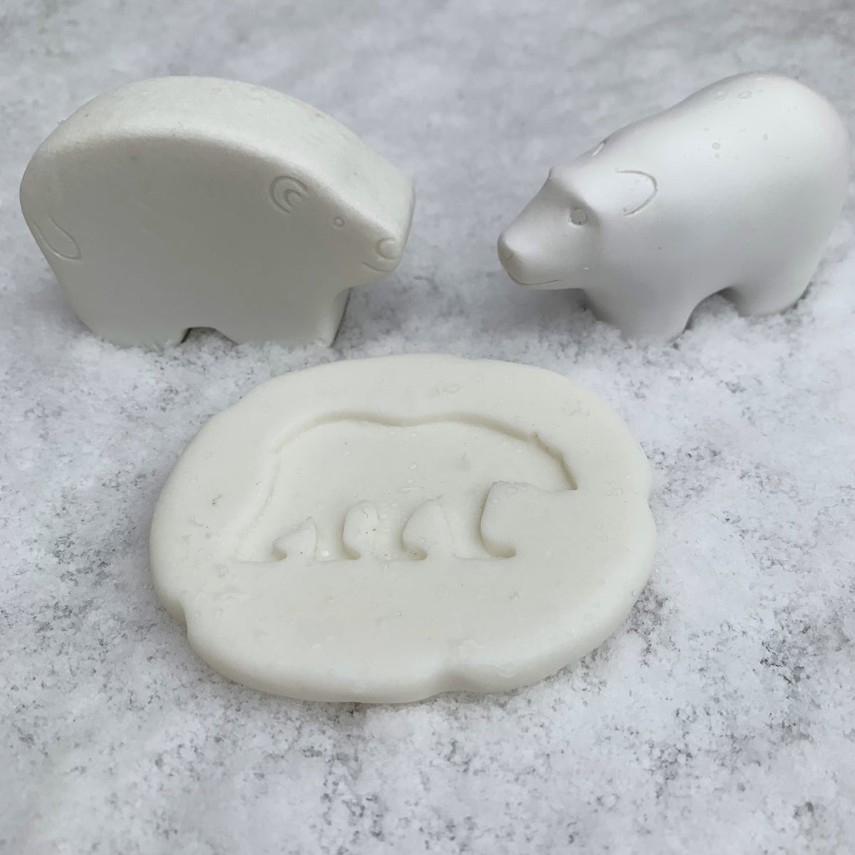 Three off-white bear-shaped items, including two small bear figurines and a larger bear-shaped mold or stamp, displayed on a snowy surface.