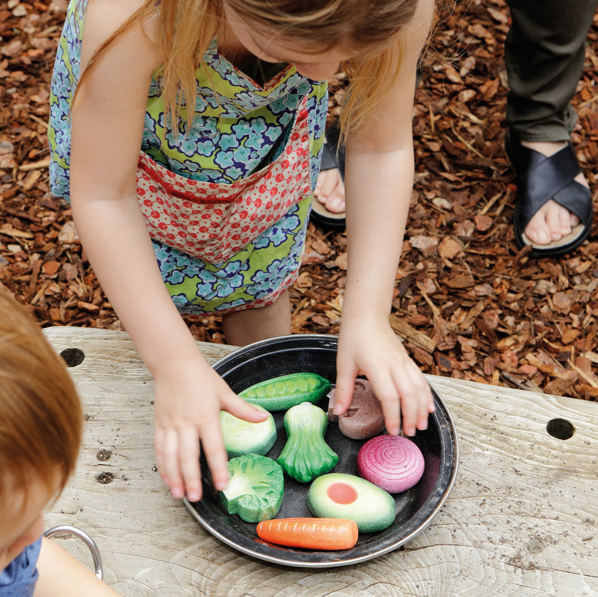 Children playing with toy play stone vegetables on a wooden surface outdoors.