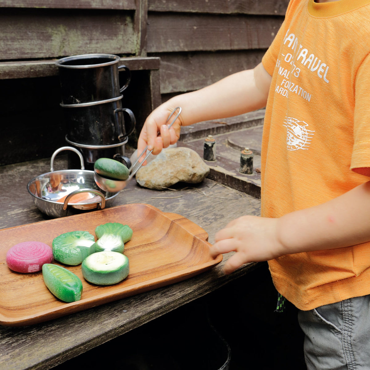 Child playing with colorful vegetable play stones on a wooden mud kitchen outdoors.