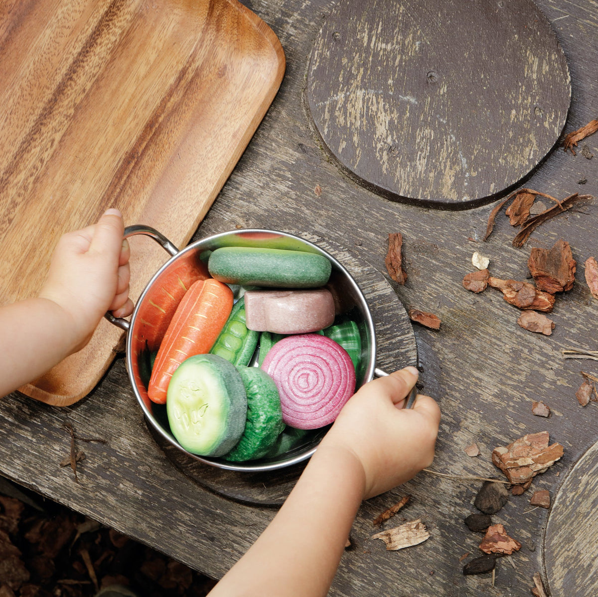 Child playing with play stone Yellow Door vegetables in a metal bowl on a wooden surface.