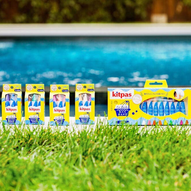 Five packages of kitpas bath crayons are lined up on a green grass poolside with a swimming pool in the background.