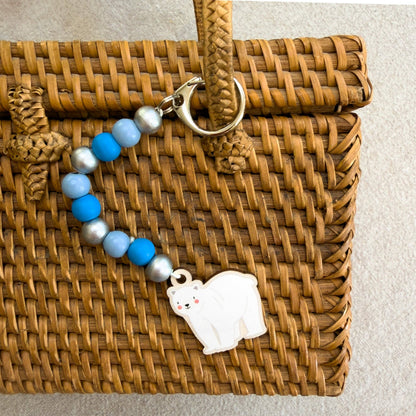 A beaded keychain or bag charm with blue and light blue beads, silver-toned beads, a gold clasp, and a white polar bear charm, displayed on a brown woven wicker bag.
