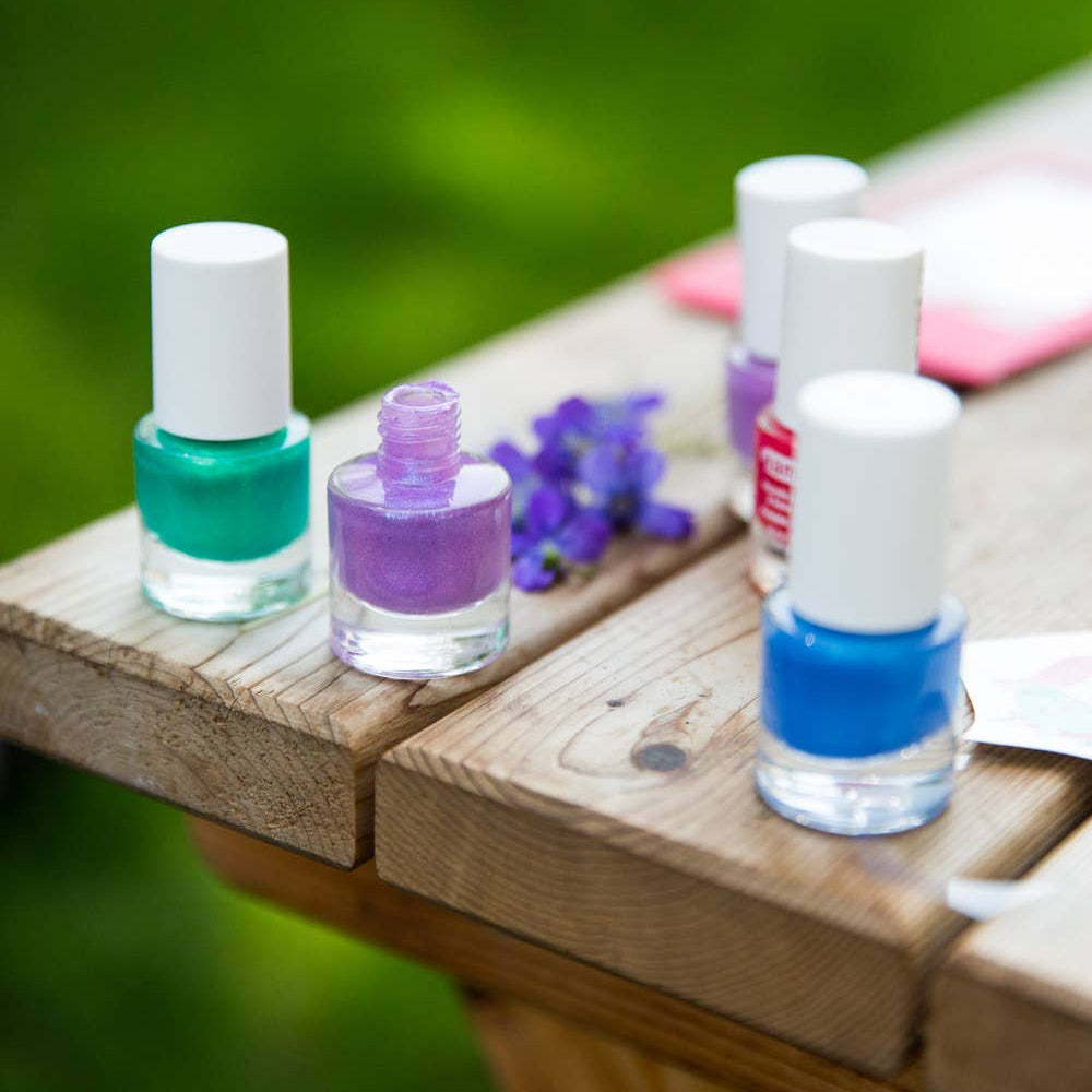 Five bottles of nail polish in green, purple, pink, red and blue, arranged on a wooden table outdoors with a blurred green background.