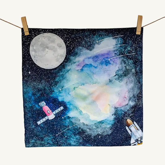 Space-themed Wonderie organic play cloth with a moon, satellite, and spacecraft on a starry background.