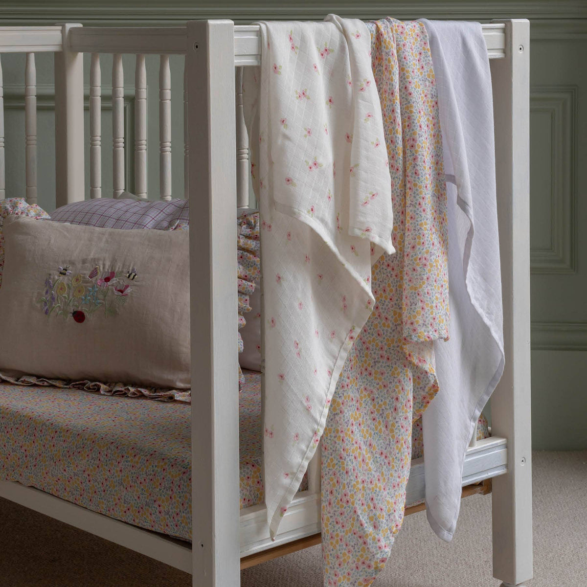 White crib with floral muslin blankets and embroidered pillow in a nursery setting