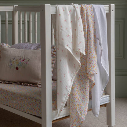 White crib with floral muslin blankets and embroidered pillow in a nursery setting