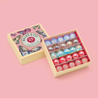 A gift box of butterfly-patterned box of marbles from Billes & Co, shown open to reveal 25 colorful spherical cards in rows of blue, silver, green, and red on a pink background.