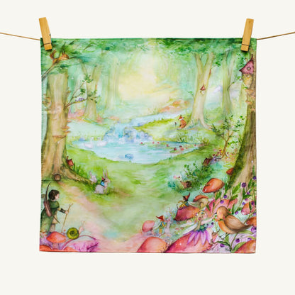 A square fabric art print or tea towel featuring a whimsical watercolor forest scene with a pond, trees, flowers, and small animals, hanging from a string with wooden clothespins on a cream fabric background.