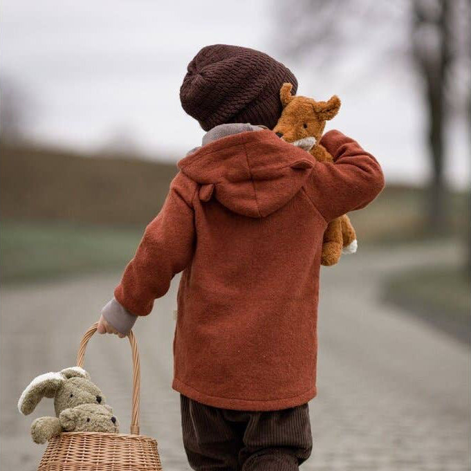 Child in a brown coat walking with a basket and stuffed animals on a cobblestone path.