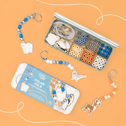 Polar Pals keyring bead kit shown with its illustrated tin packaging and an open tin displaying the bead compartments and keyring components, arranged on a peach background with decorative rope lines.