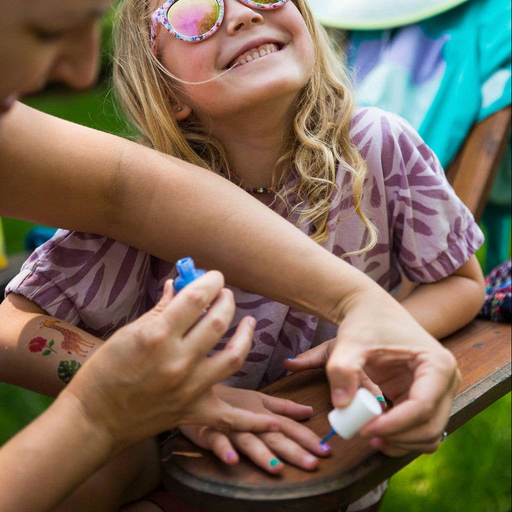 A young girl wearing pink mirrored sunglasses and a purple patterned shirt smiles while having her fingernails painted by an adult outdoors on a wooden chair.