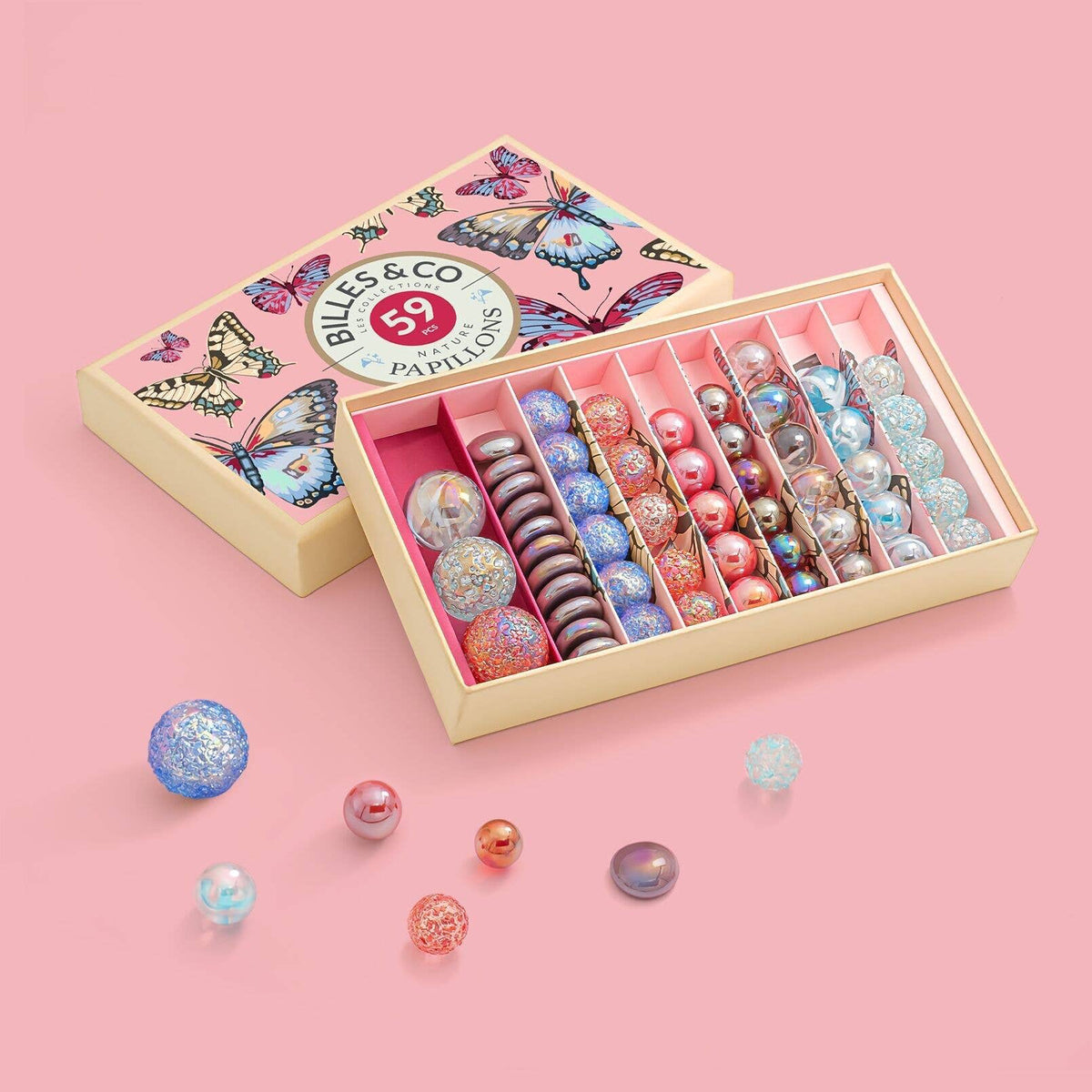 An open decorative pink butterfly gift box of glass marbles from Billes & Co. shown alongside its illustrated lid on a solid pink background, with several loose marbles scattered in front.