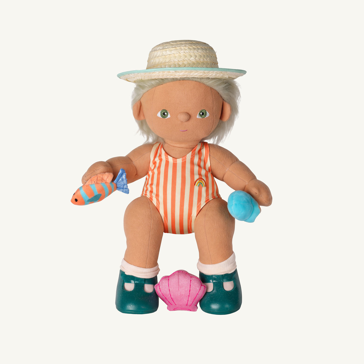 A dinkum doll with white hair wearing a straw hat, orange and white striped swimsuit, teal boots, and sitting on a pink seashell, holding a fish toy and blue ice cream toy, on a light off-white background.