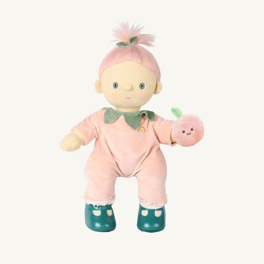 A plush dinkum doll with pale skin and green eyes, wearing a pink velvet one-piece outfit with a green collar, teal shoes, and holding a small pink plush peach, isolated on a white background.