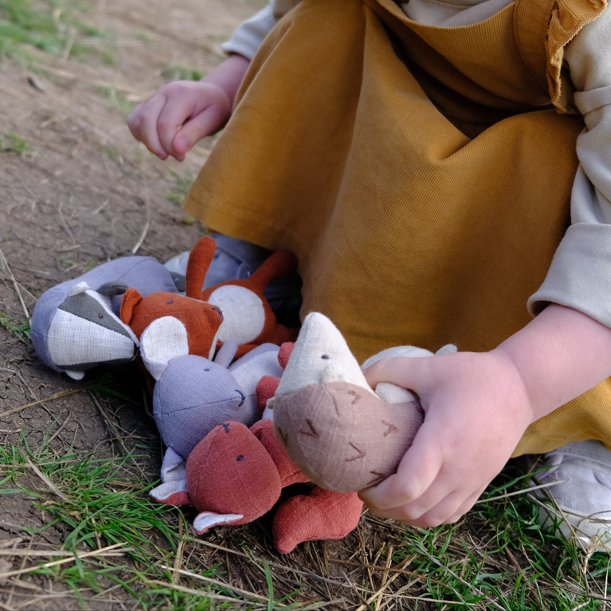 A young child with blonde hair sits outdoors on grass and dirt, holding a plush stuffed animal among several other Nutshell soft toys.