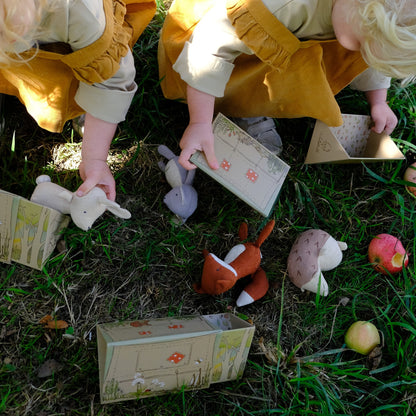 Two toddlers in cream and mustard yellow outfits play outdoors on grass, interacting with a Nutshell toy set that includes printed cardboard tents, a plush rabbit, a fox, and a hedgehog.