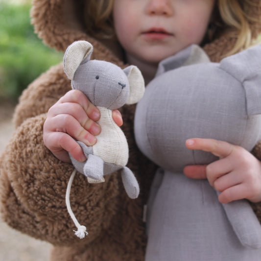 Child holding two plush toys, one small gray mouse and one large gray mouse, outdoors.