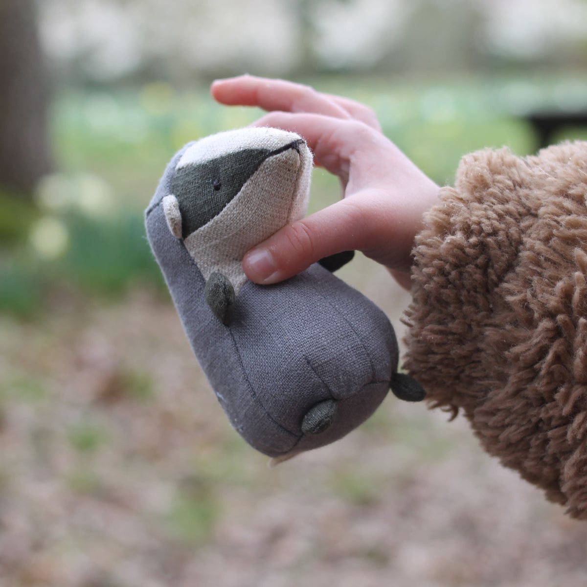 Person holding a small gray plush Nutshell badger outdoors with a blurred natural background
