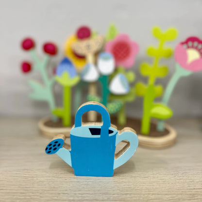 Blue watering can toy with a blurred background of colorful flowers and plants.