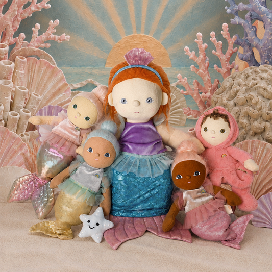 A group of plush mermaid dolls arranged on a sandy surface with a painted ocean backdrop featuring sun rays, coral, shells, and birds. The central doll has orange hair, a purple top, and a blue tail, while surrounding dolls wear pink, blue, and brown outfits.