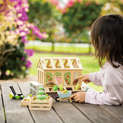 Child playing with a toy greenhouse and accessories on a wooden table outdoors.