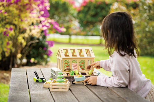Child playing with a toy greenhouse and accessories on a wooden table outdoors.