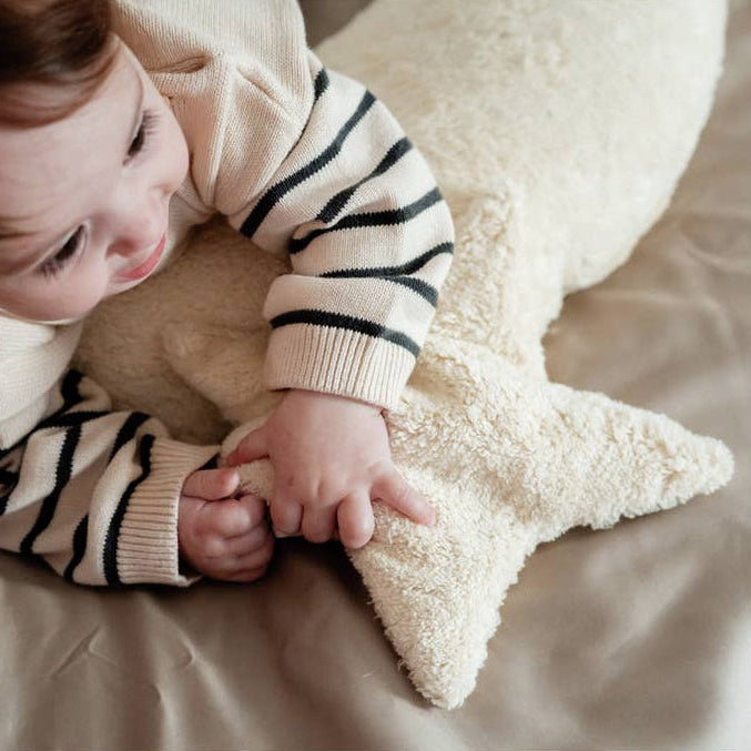 A baby wearing a cream knit sweater with black stripes lies on a beige bed, holding a fluffy cream star-shaped pillow.