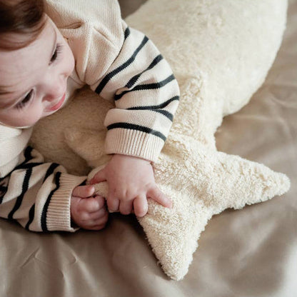 A baby wearing a cream knit sweater with black stripes lies on a beige bed, holding a fluffy cream star-shaped pillow.
