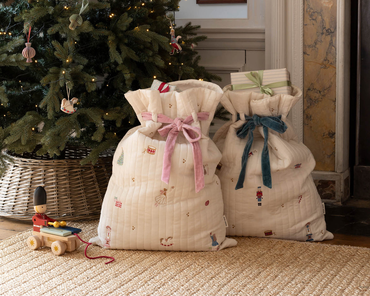 Two quilted cream fabric Avery Row christmas stocking sacks with festive prints and ribbon bows, placed on a woven rug in front of a decorated Christmas tree and fireplace.