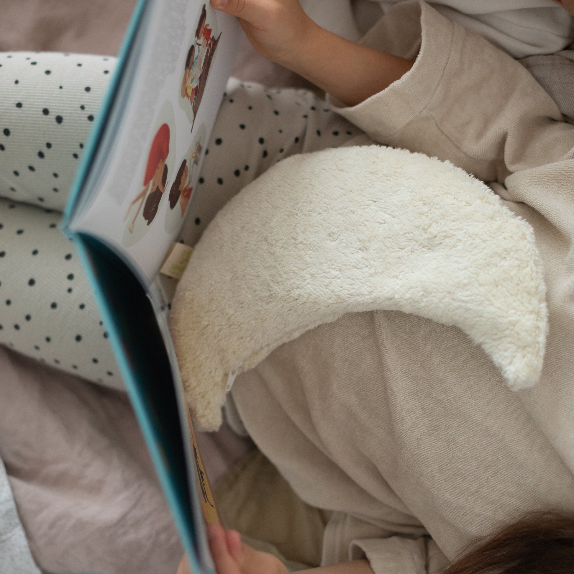A cozy lifestyle shot of a child reading a picture book, nestled in soft beige pajamas and surrounded by plush toys and a cream crescent moon pillow on a bed.
