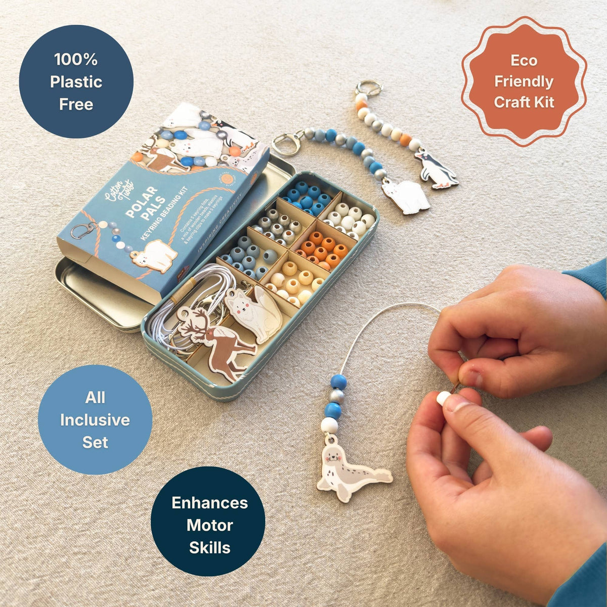 A children's eco-friendly craft kit shown on a beige carpet, featuring a metal tin with bead compartments, a beaded animal charm, and a pair of hands actively assembling a bead and charm. The image includes text overlays highlighting product features like '100% Plastic Free', 'Eco Friendly Craft Kit', 'All Inclusive Set', and 'Enhances Motor Skills'.