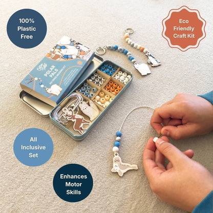 A children's eco-friendly craft kit shown on a beige carpet, featuring a metal tin with bead compartments, a beaded animal charm, and a pair of hands actively assembling a bead and charm. The image includes text overlays highlighting product features like '100% Plastic Free', 'Eco Friendly Craft Kit', 'All Inclusive Set', and 'Enhances Motor Skills'.