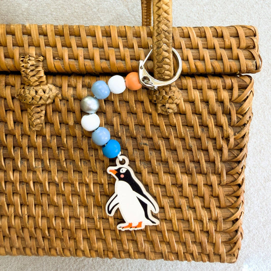 A beaded keychain with a penguin charm hanging from a silver clasp, displayed on a brown woven rattan bag against a light gray background.