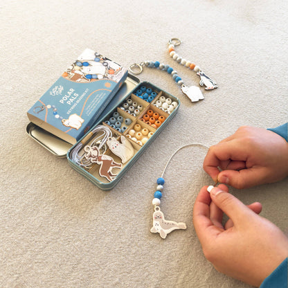 A person's hands are shown assembling a beaded keychain charm from a Cotton Twist kit on a beige carpet. The kit includes a blue and white illustrated box, an open metal organizer filled with beads and charms, and a completed keychain with blue beads, white beads, and a white polar bear charm.