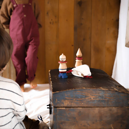 Child in maroon overalls standing behind a wooden chest with small figurines on top, in a room with wooden walls.